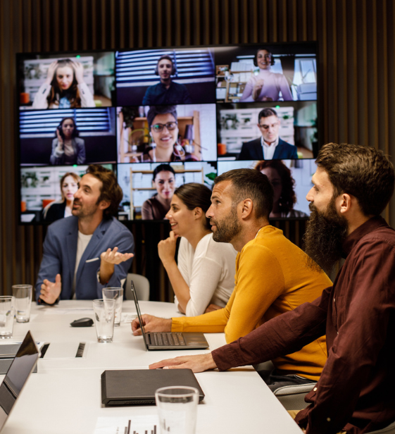 Hybrid team in a conference room engaged in a video call with global colleagues — reflecting the complexity of supporting a dispersed workforce.