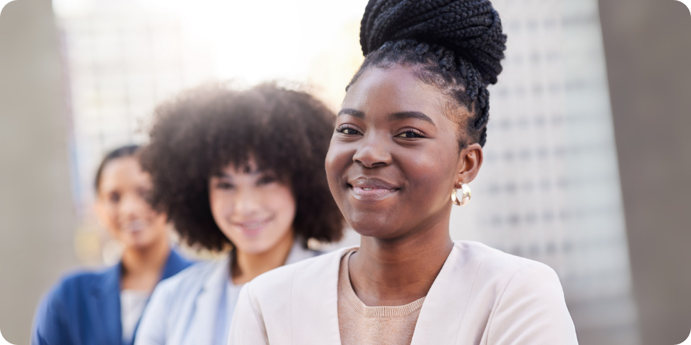 Confident young woman smiling with supportive colleagues behind her — representing a new approach to helping women thrive at work. Confident young woman smiling with supportive colleagues behind her — representing a new approach to helping women thrive at work.